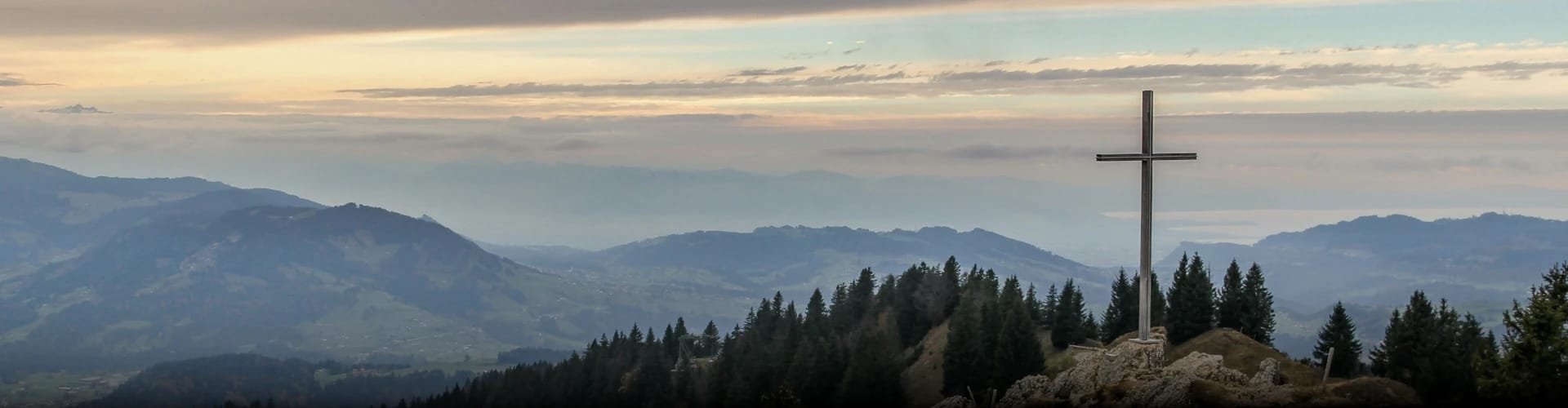 A cross on the top of a hill with foothills in the background, all covered with pine trees.
